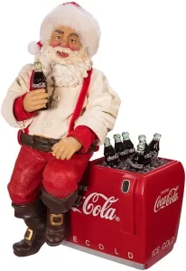 Adorno navideño de Santa Claus con gorro rojo, camisa color perla con tirantes rojos y pantalon rojo y botas negras sentado en una hielera roja con refrescos de la marca cocacola.